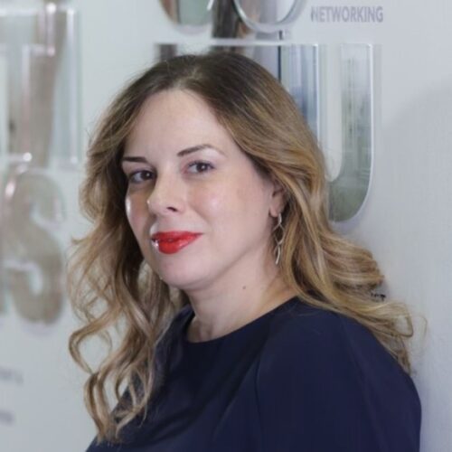 Ένα άτομο με μακριά, κυματιστά μαλλιά και κόκκινο κραγιόν φοράει σκούρο τοπ. Στέκονται μπροστά σε έναν τοίχο με αχνά, θολά γράμματα στο βάθος, δίνοντας μια επαγγελματική και γεμάτη αυτοπεποίθηση εμφάνιση.